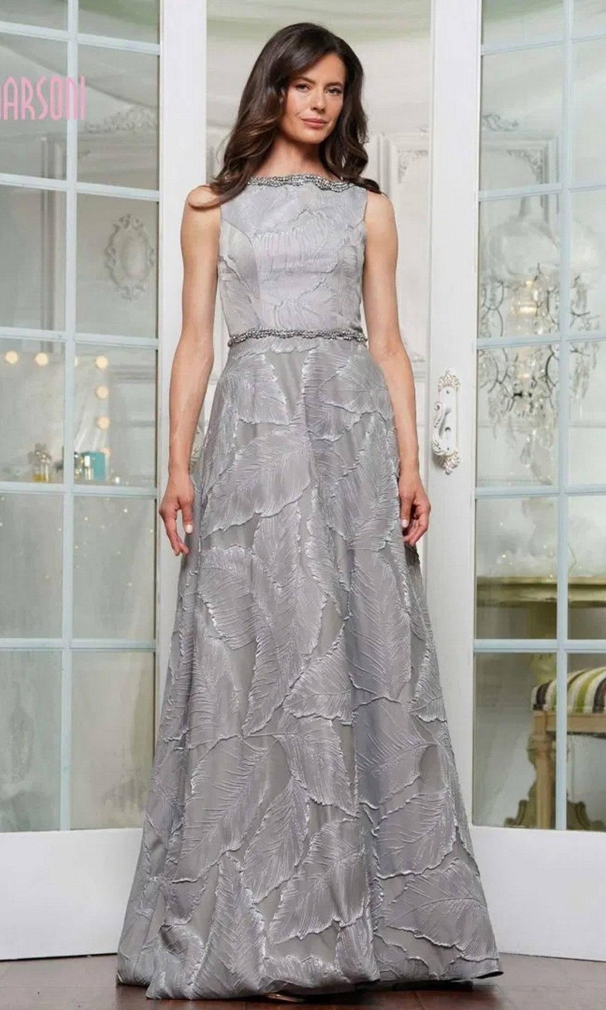 A woman poses indoors in front of glass-paneled doors, wearing the Marsoni MV1224 long formal dress in charcoal—a sleeveless gown with a subtle jacquard print and belted waist.